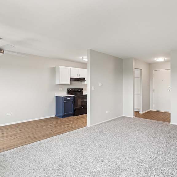 a spacious room with a grey carpet and kitchen in the background