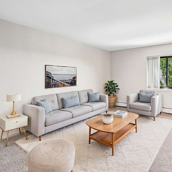 a living room with a grey couch, a wooden coffee table, a white rug, a white wall, a window with a view of trees, and a painting hanging on the wall.
