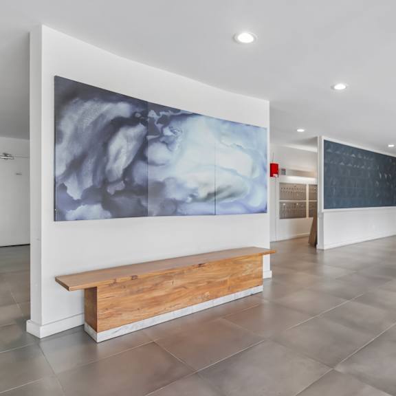 a large grey and white painting hangs on a white wall above a wooden bench in a spacious room.