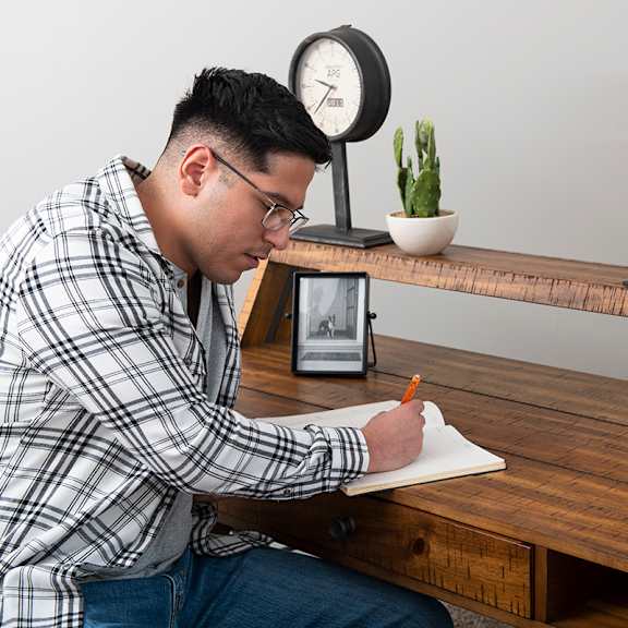 a man is writing on a piece of paper while sitting at a wooden desk with a clock, books, and a small potted plant on the shelf behind him.