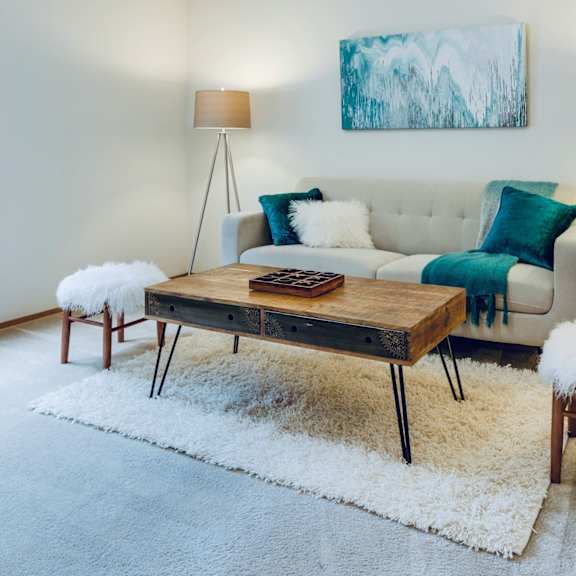 a white couch with blue pillows and a wooden coffee table with a white fluffy stool in front of it.