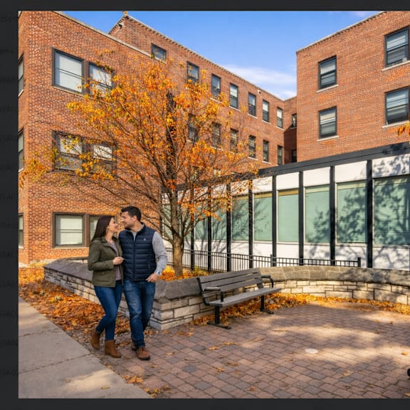 a couple is standing on a brick patio in front of a brick building with a tree with orange leaves.