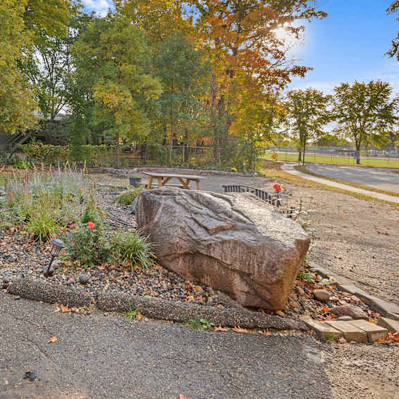 a large rock sits in the middle of a gravel area with a grassy area and trees in the background.