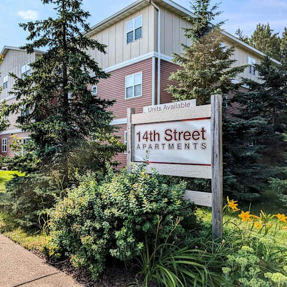 a sign for 14th street apartments is in front of a building with a well-kept lawn and trees.