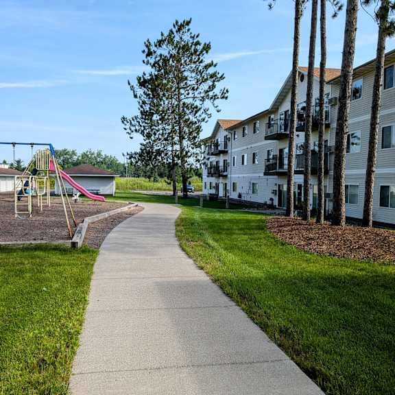 a playground with a slide and swings is surrounded by a grassy area and a sidewalk.