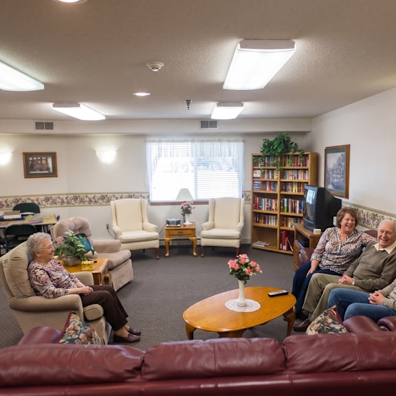 a group of people are sitting in a cozy room with a red couch, a coffee table, and a bookshelf.