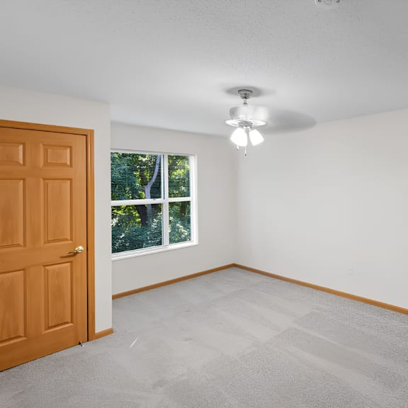 a room with a carpeted floor, a ceiling fan, and a window with a view of trees outside.
