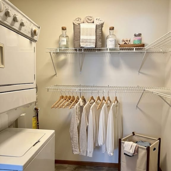a laundry room with a washer and dryer, shelves with towels and other items, and clothes hanging on a rack.