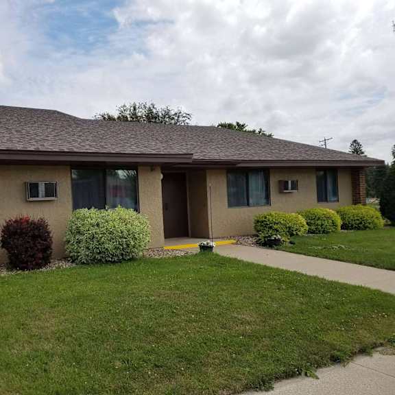 a house with a brown roof and beige walls with bushes in front.