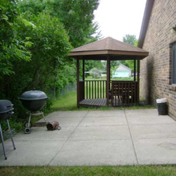 a gazebo is attached to a house and has a grill and a chair in front of it.