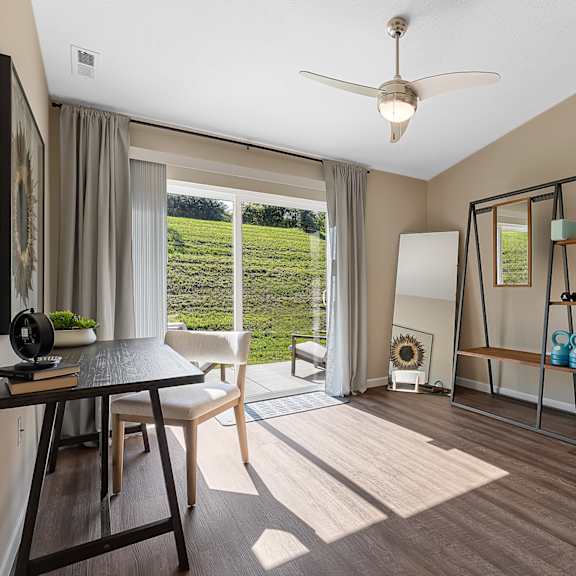 a room with a ceiling fan, a table with a chair, a potted plant, and a view of a green field through the window.
