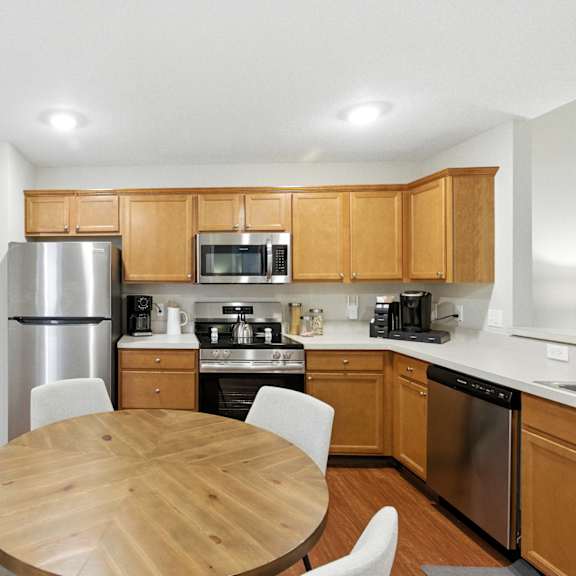 a kitchen with a table and chairs in the foreground and a refrigerator, microwave, oven, and sink in the background.