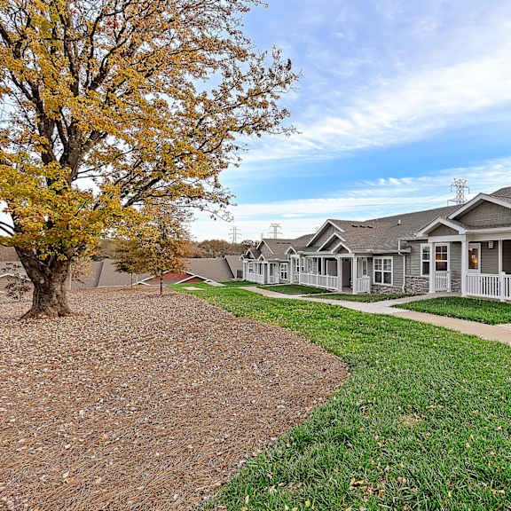 a house with a grey roof and a white fence is surrounded by a grassy area and a tree with yellow leaves.