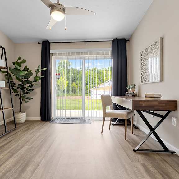 a room with a sliding glass door leading to a backyard, a desk with a chair, and a shelf with decorative items.