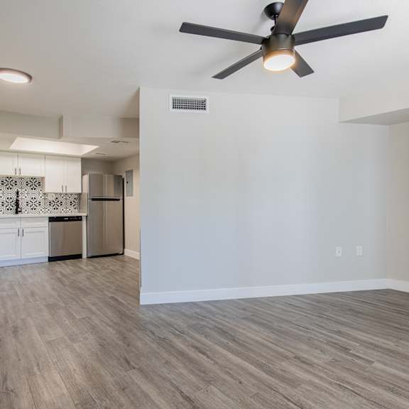 an empty living room with a ceiling fan and a kitchen in the background