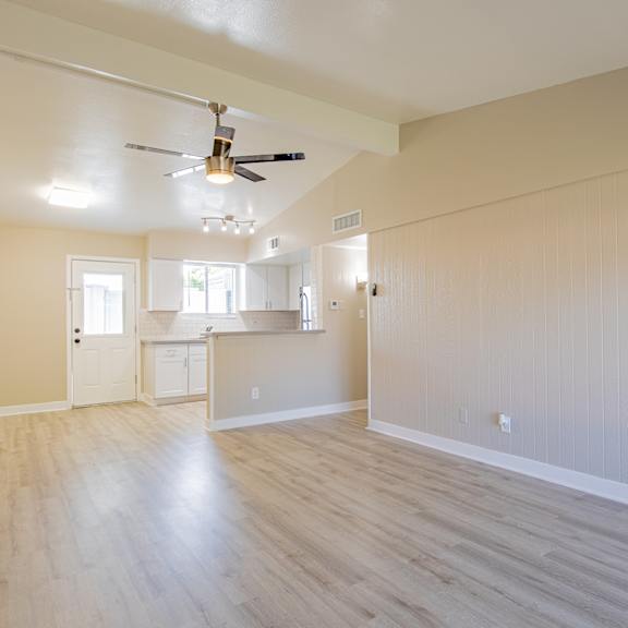 an empty living room with a ceiling fan and a kitchen in the background