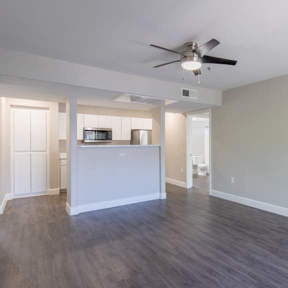 an empty living room with a ceiling fan and a kitchen in the background