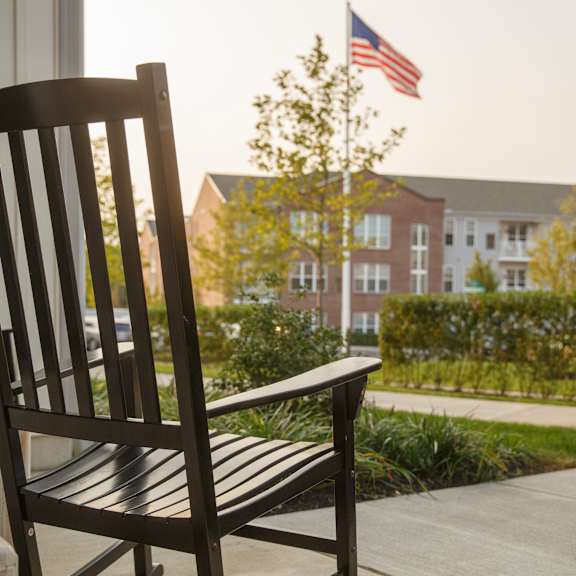 a wooden rocking chair sits on a porch with a building and an american flag in the background.