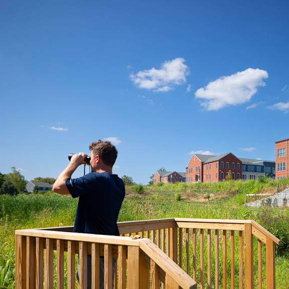 a man is standing on a wooden deck and looking through binoculars at a large red building.