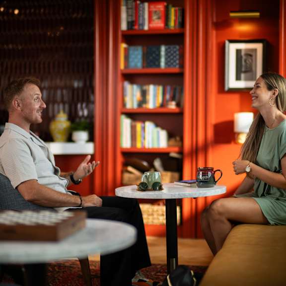 a man and a woman are having a conversation in a cozy room with a bookshelf in the background.