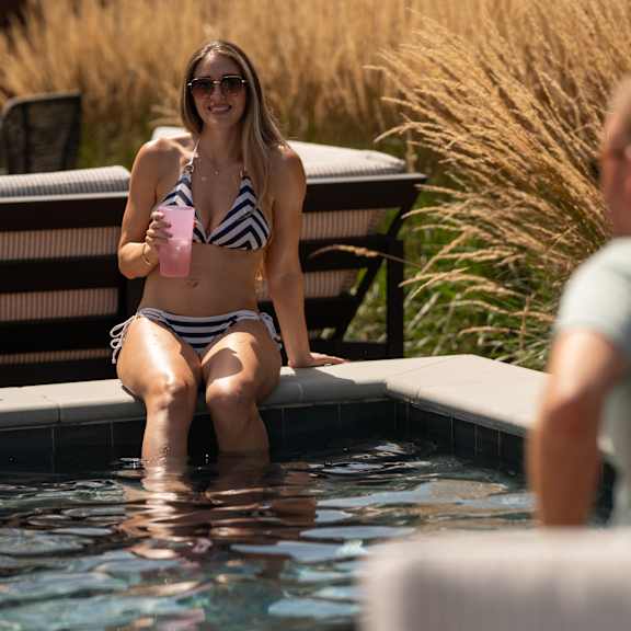 a woman in a striped bikini sits on the edge of a pool holding a drink.