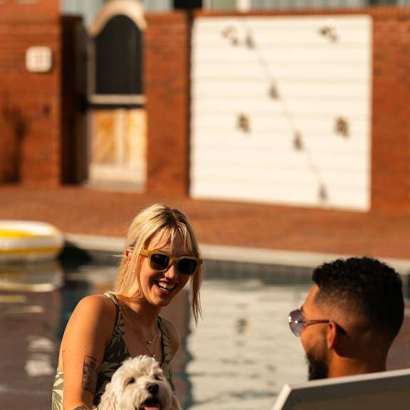a woman and a man are sitting by a pool with a dog, looking at a laptop.