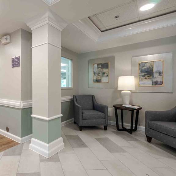 a hallway with a grey sofa, armchair and a small table with a white lamp on it.