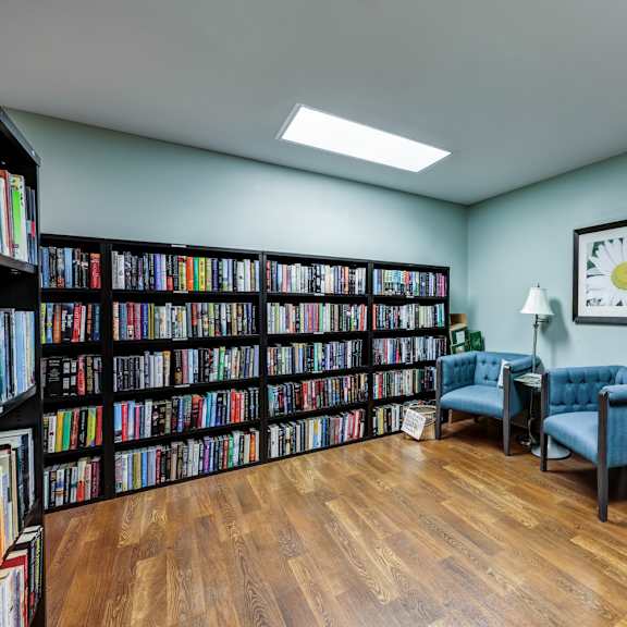 a room full of books with a blue chair and a lamp.