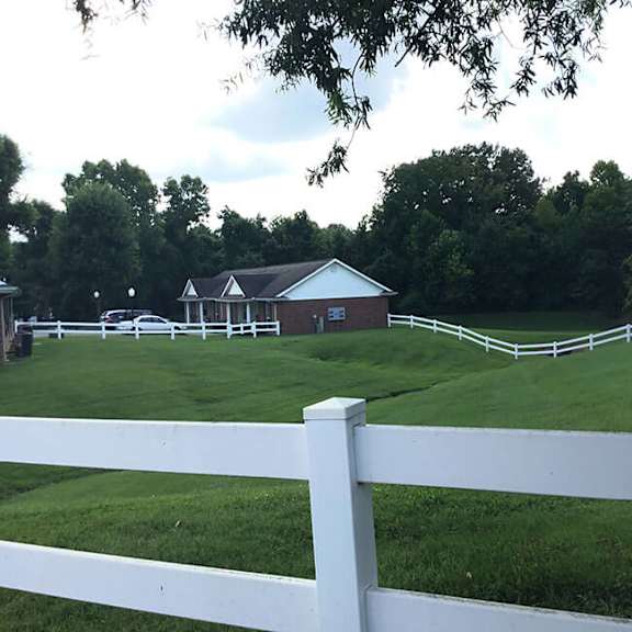 a white fence in front of a green field with a building in the background.