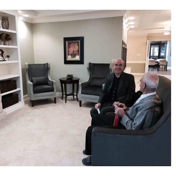 two men are sitting in a room with a fireplace and a painting on the wall.