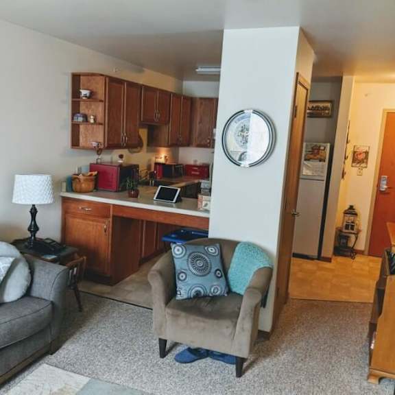 a living room with a grey couch, a chair, and a kitchen area in the background.