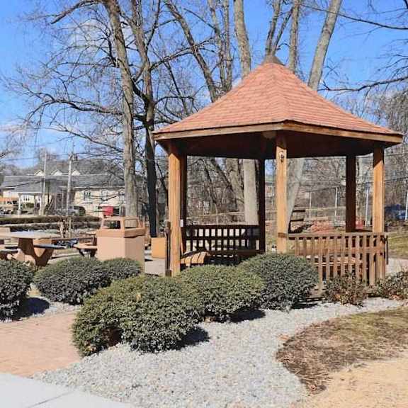 a gazebo with a brown roof is surrounded by bushes and gravel.
