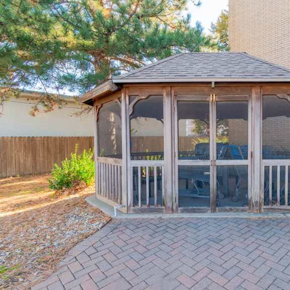 a gazebo with a brown roof and glass panels is surrounded by a brick walkway.