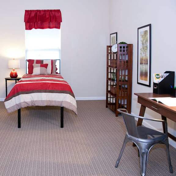 a bedroom with a bed, desk, chair, and a window with red curtains.