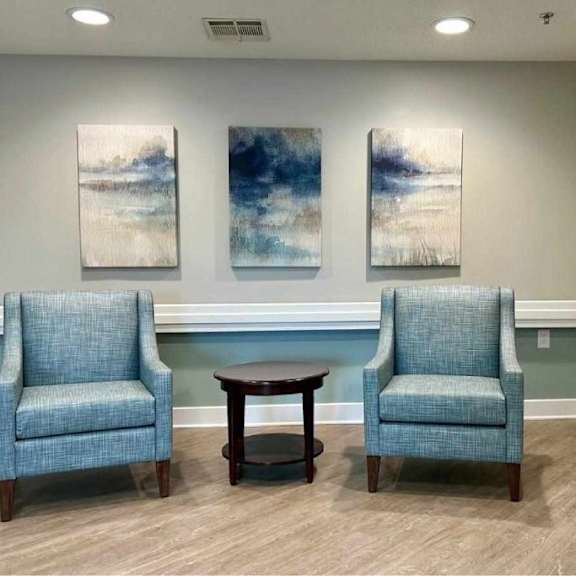 two blue chairs and a table in a room with two paintings on the wall.