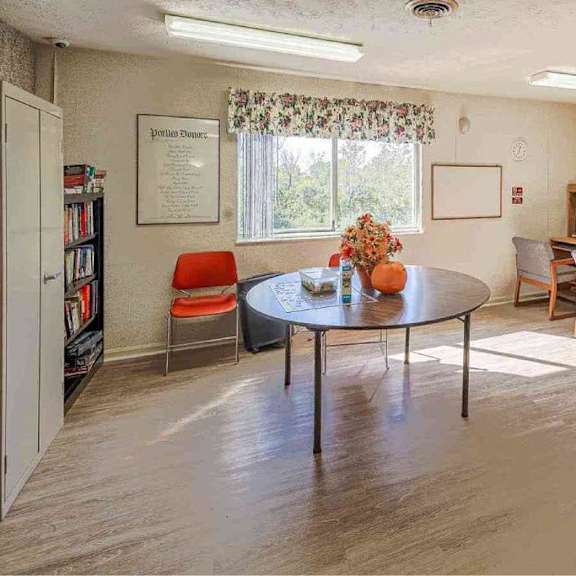 a living room with a round table and chairs, a bookshelf, and a window with curtains.
