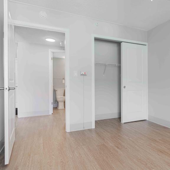 a bedroom with white walls, wood floors, double door closet and a view into the hallway and restroom.