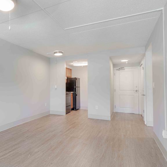 the living area with an overhead ceiling fan with light with white walls, wood floors looking into the kitchen and the main entry door.