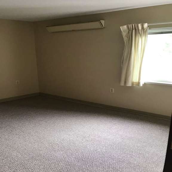 a room with a carpeted floor, a window with curtains, and an air conditioner on the wall.