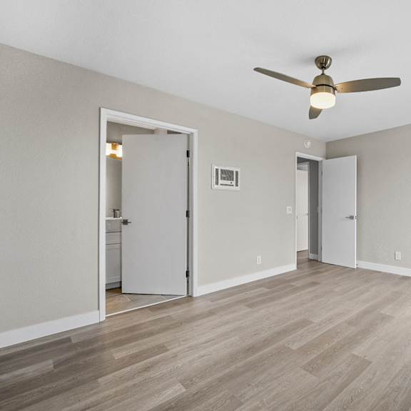 a room with a ceiling fan and light fixture, hardwood floors, and white walls with a doorway leading to another room at tamarack sands apartment homes, carlsbad, california