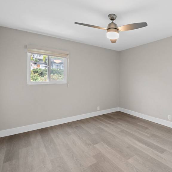 a room with a ceiling fan and light, hardwood floors, and a window with a view of a building.