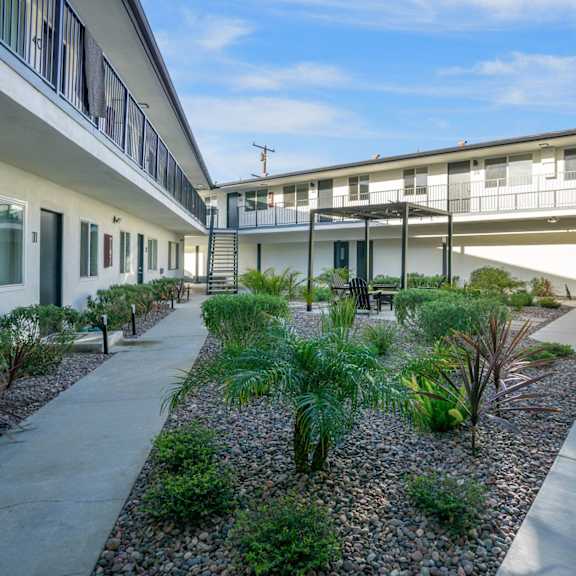 a concrete walkway leads between two buildings with a landscaped area in the middle. at torrance gateway apartments, california, 90501