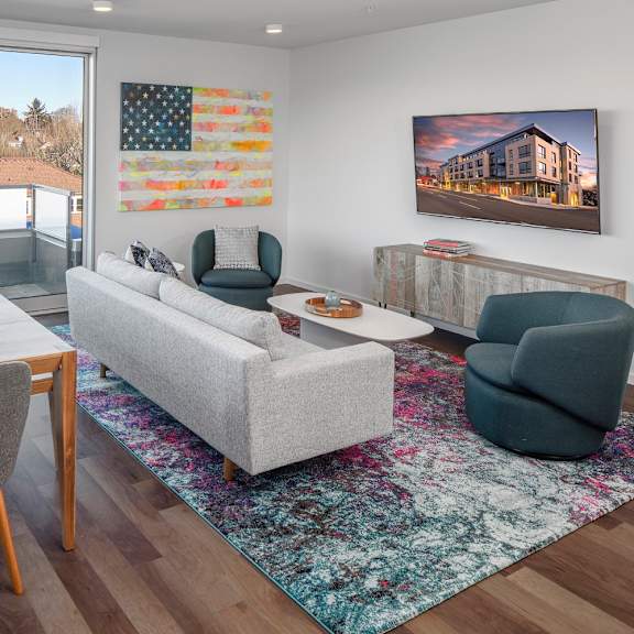 a living room with a grey couch, a wooden dining table and a painting of a building on the wall.