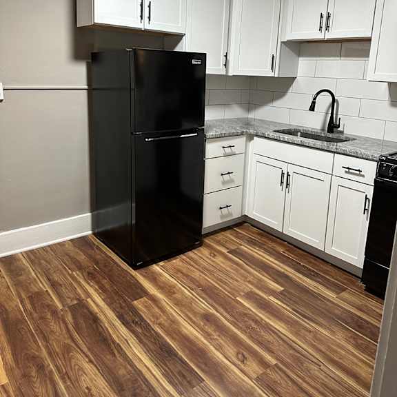a black refrigerator in a kitchen with white cabinets and a wooden floor.