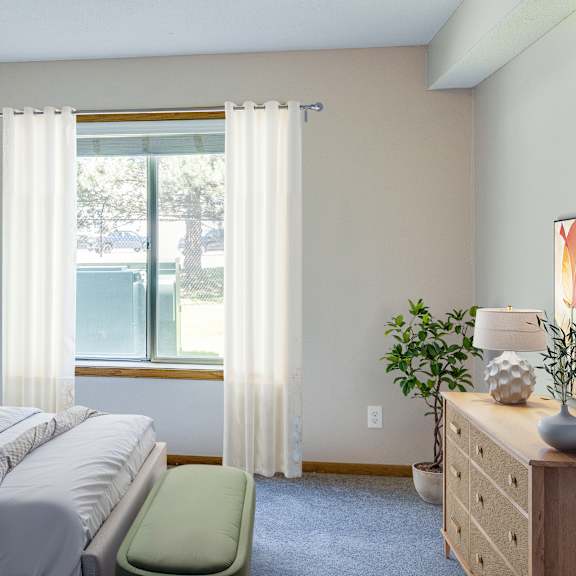 a bedroom with a bed, a chair, a nightstand, and a window with curtains. at crosby pointe apartments, st. paul , mn, 55116-3244