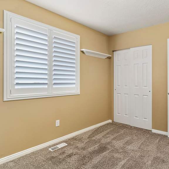 a room with a carpeted floor, two white doors, and a window with white shutters.