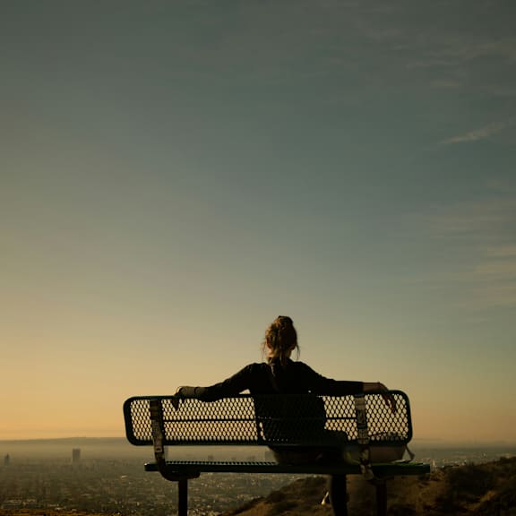 a person is sitting on a bench overlooking a cityscape at sunset.