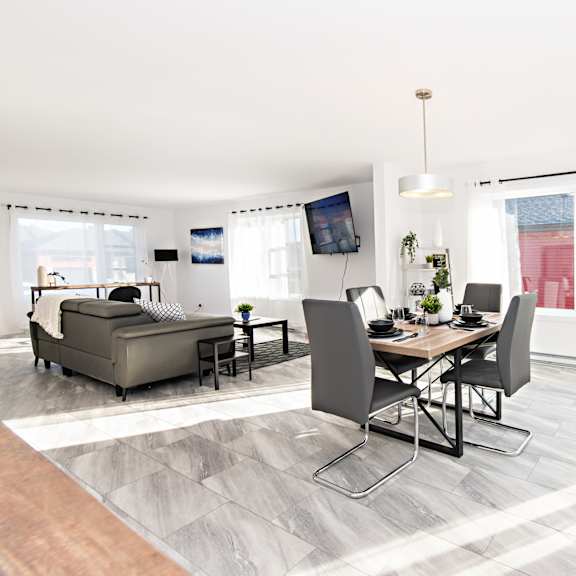 a modern living room with a grey sofa, a wooden dining table with four chairs, and a television mounted on the wall.