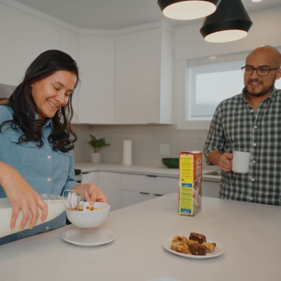 a woman pours milk into a bowl while a man stands behind her holding a cup.
