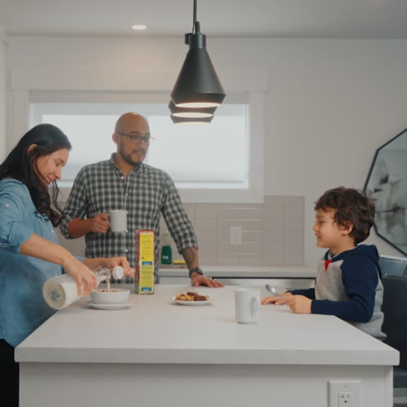 a family of three is gathered around a kitchen island, with a young boy sitting at the table and two adults standing nearby.